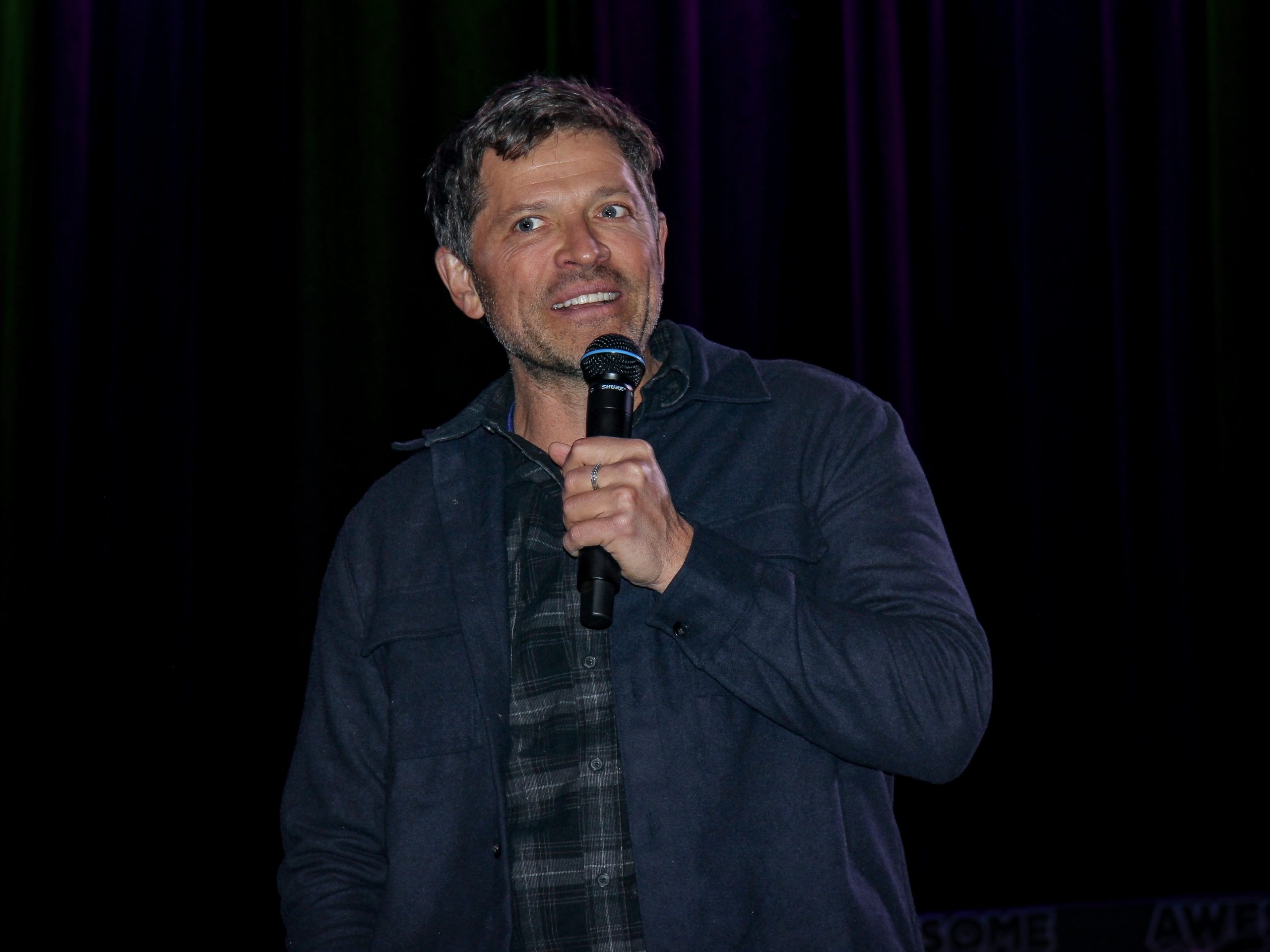 Misha Collins holds a microphone as he addresses the crowd from the stage during his Awesome Con panel.